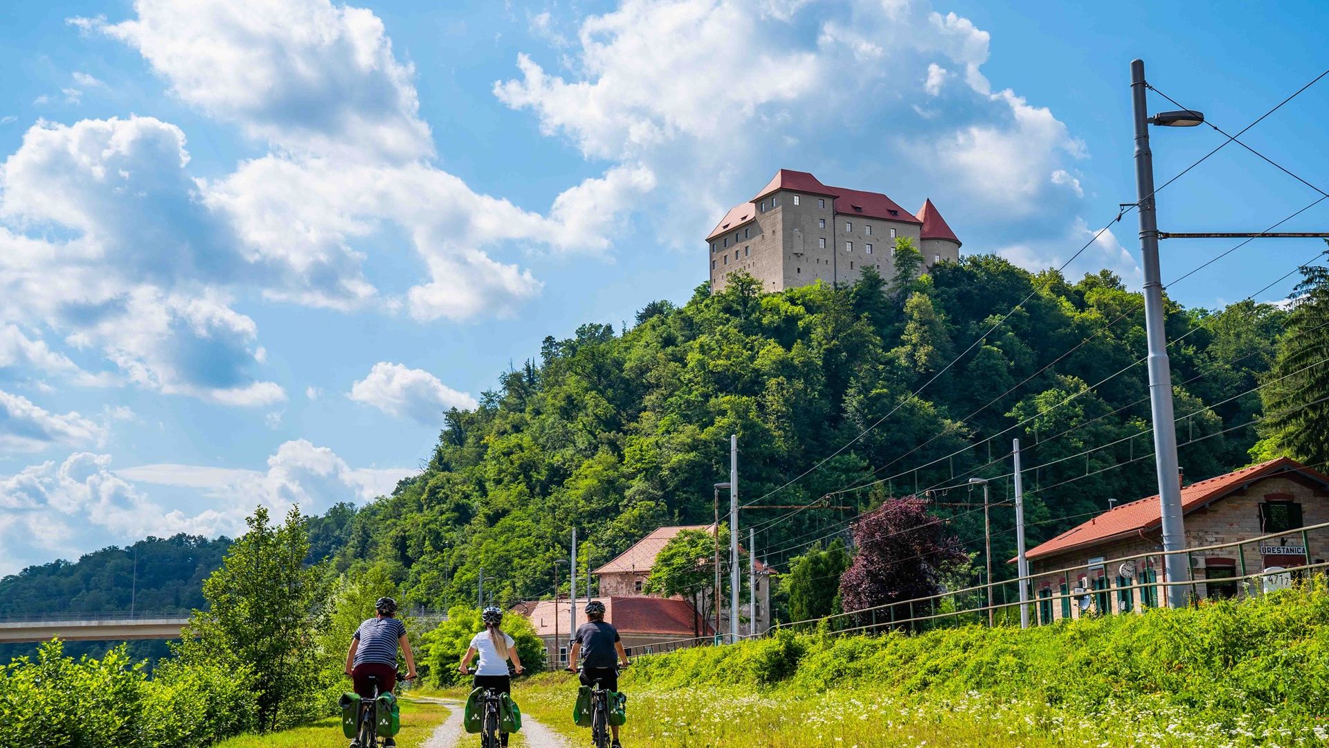 Fotografija treh ljudi, ki kolesarijo po travnati poti, nad njimi pa je grad.